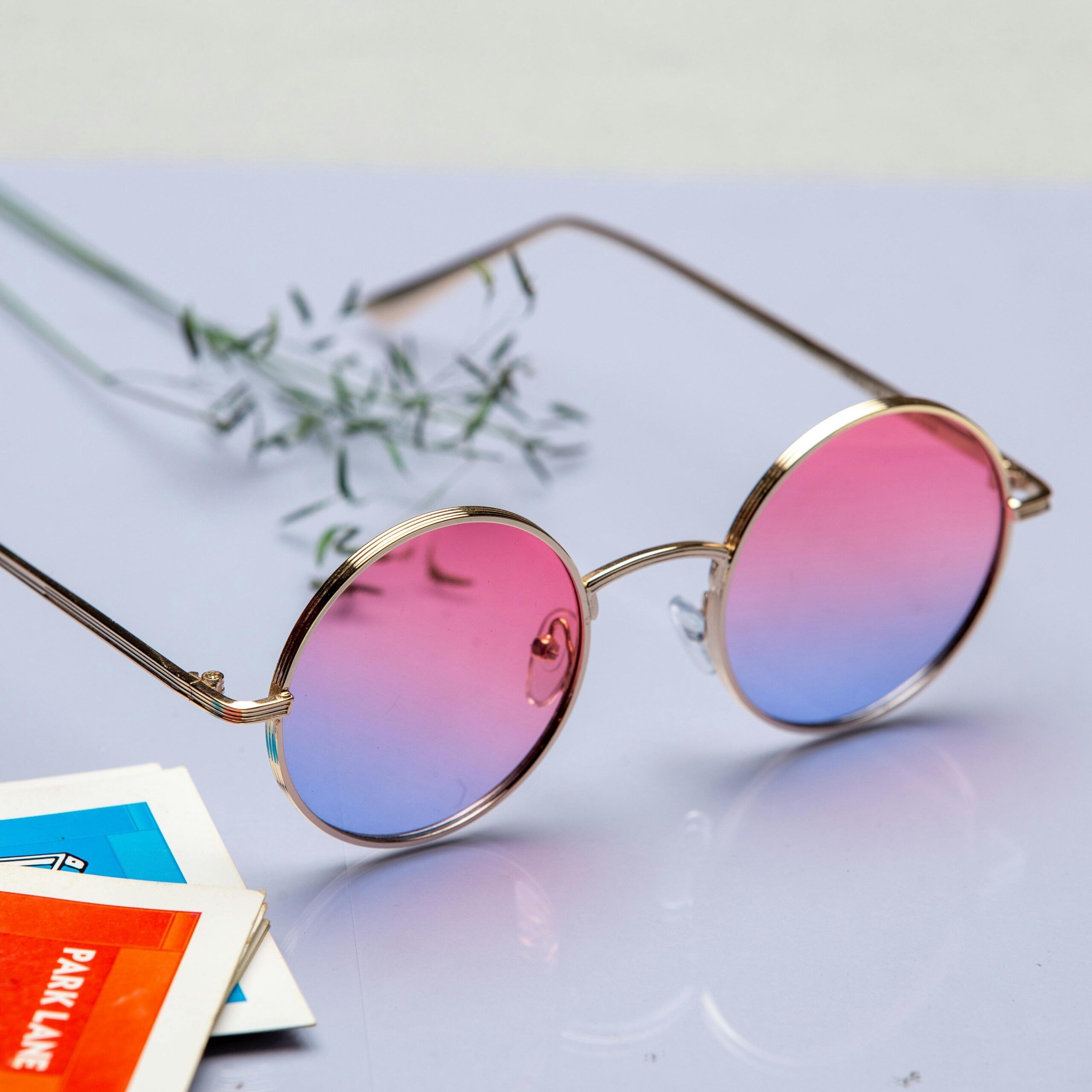 Round sunglasses with pink to blue gradient lenses on a pastel surface, evoking a trendy vibe.