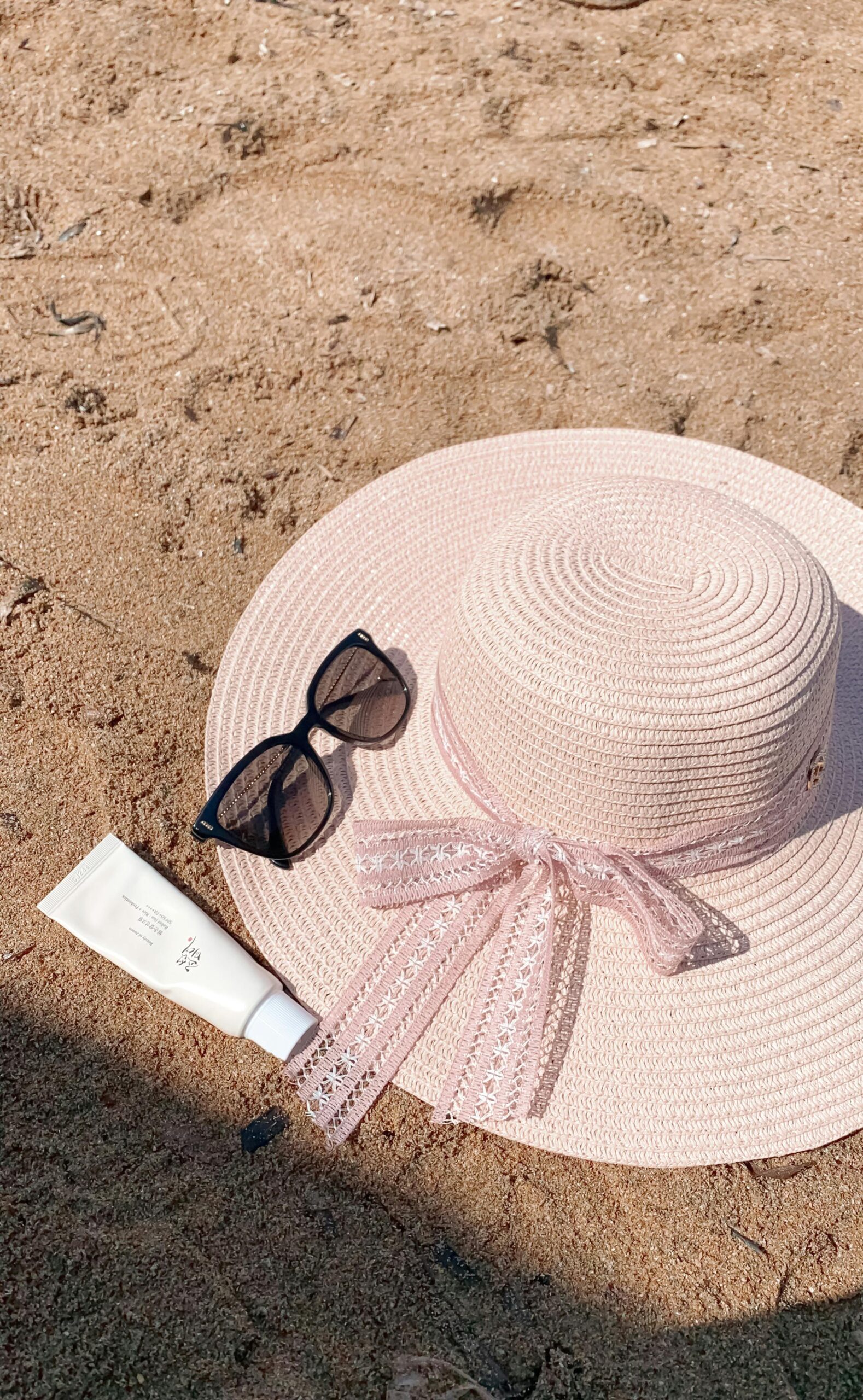 Stylish pink sun hat with sunglasses and sunscreen on a sandy beach, perfect for summer relaxation.