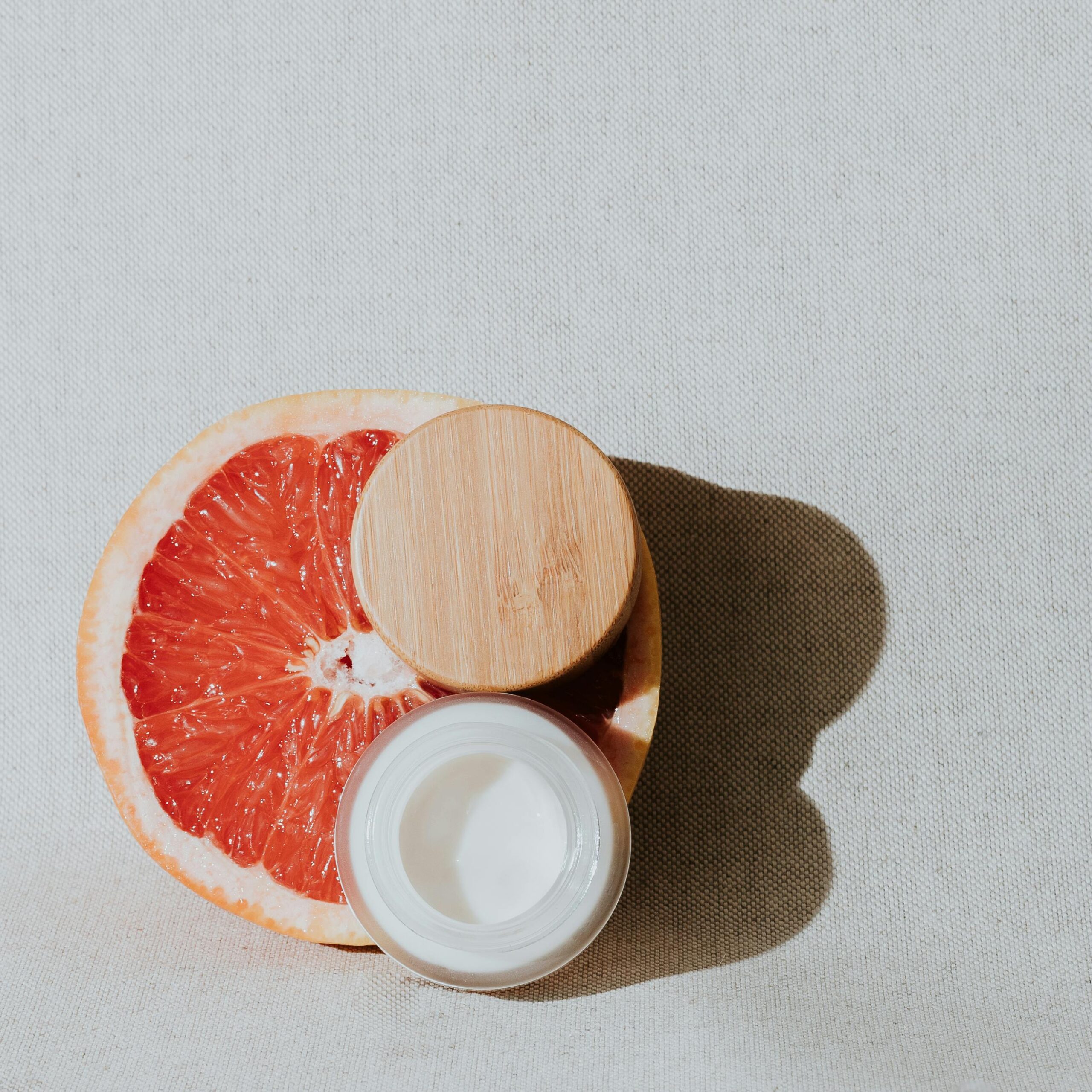 Flat lay of a grapefruit half and cream jar with bamboo lid on a neutral canvas background.