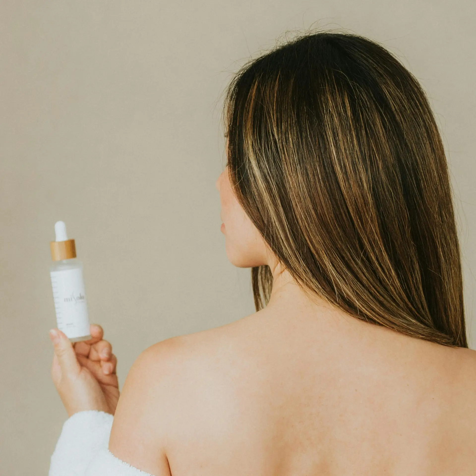 Minimalist studio shot of a woman holding a skincare serum against a beige wall, emphasizing beauty and self-care.