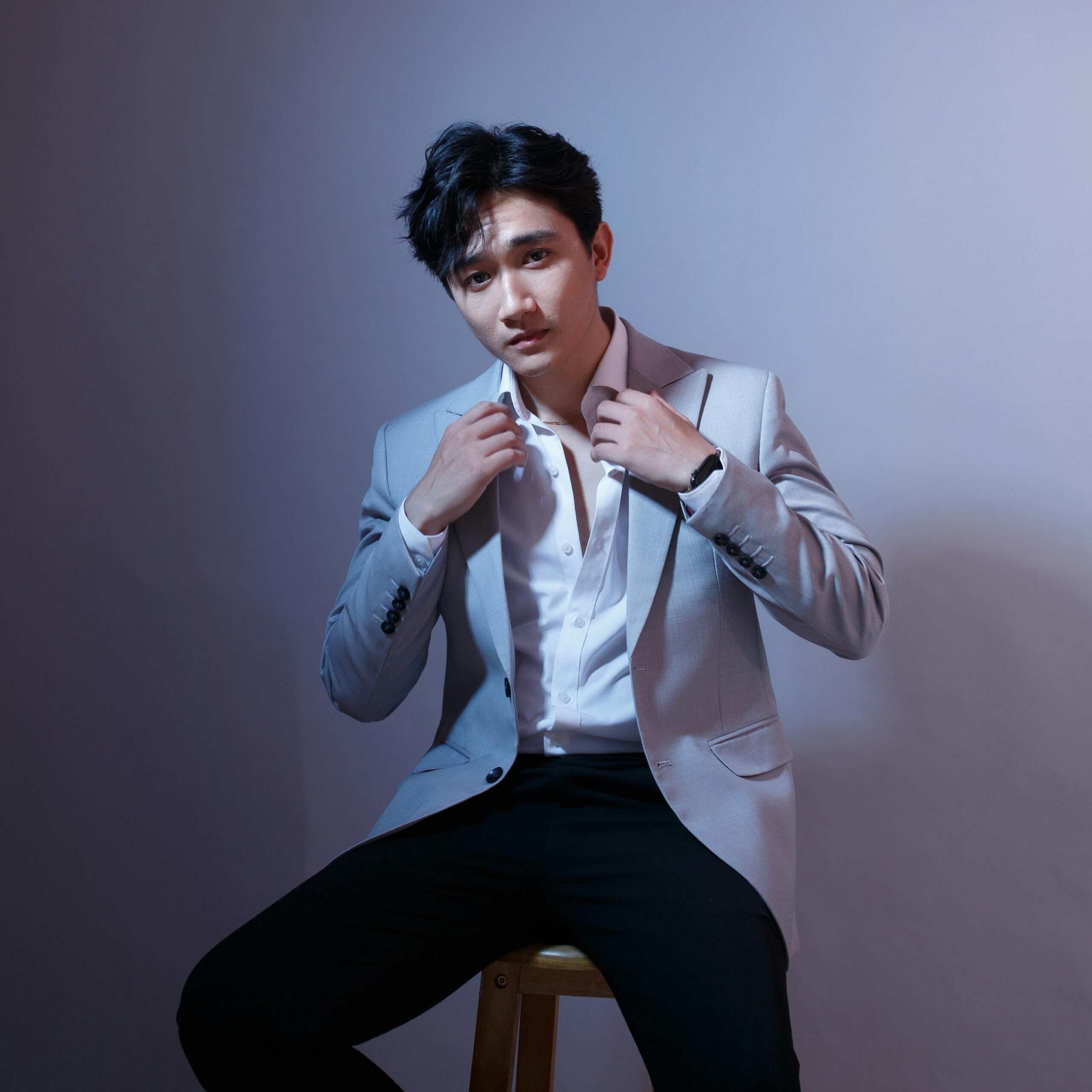 A stylish man in a suit poses on a stool against a gray background, exuding confidence and elegance.