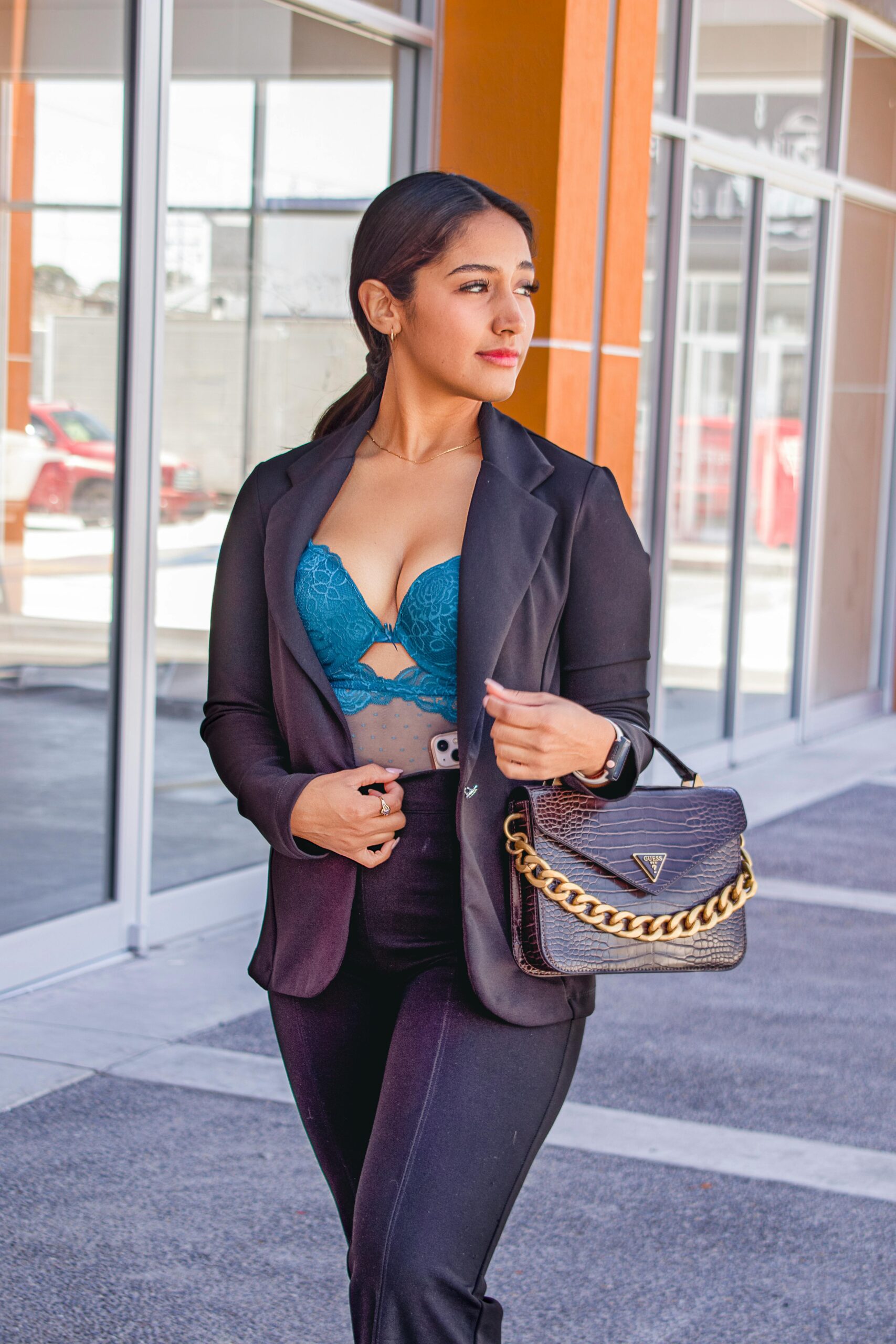 Elegant woman walking outdoors in a black suit and blue lingerie, holding a purse.