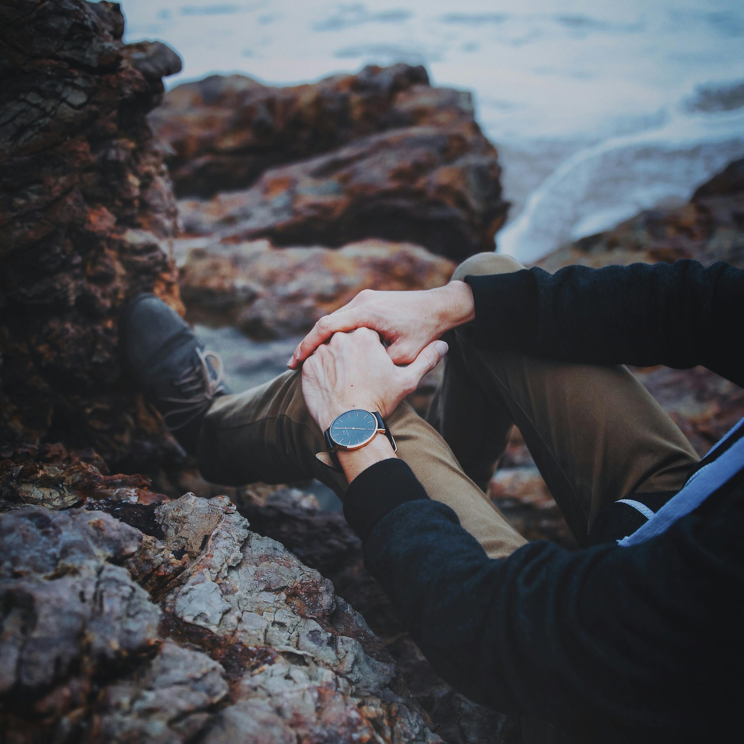 A person sitting on a rocky shore enjoying the sea view with hands clasped, wearing a wristwatch.
