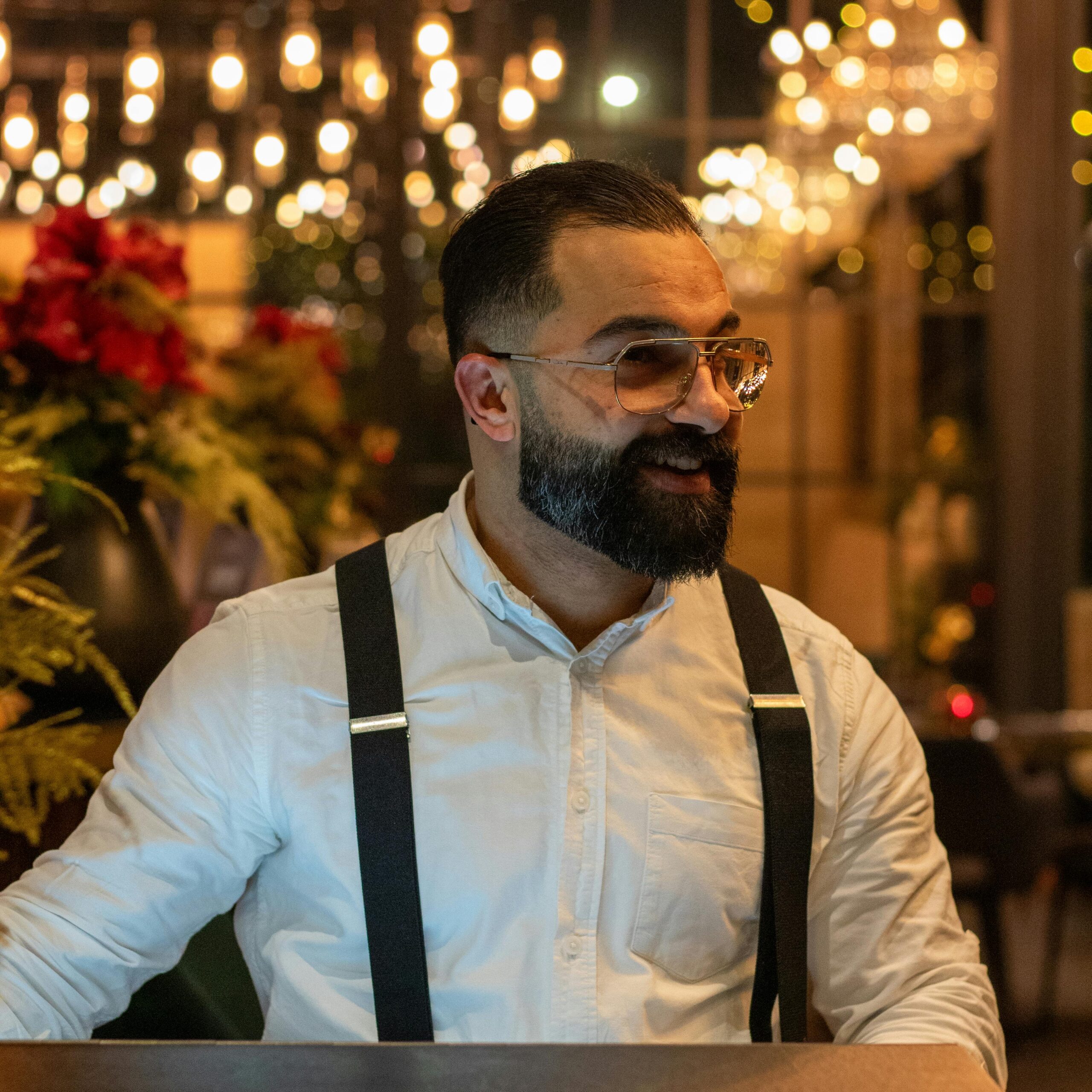 A smiling bearded man in a white shirt and suspenders, enjoying a festive indoor environment.