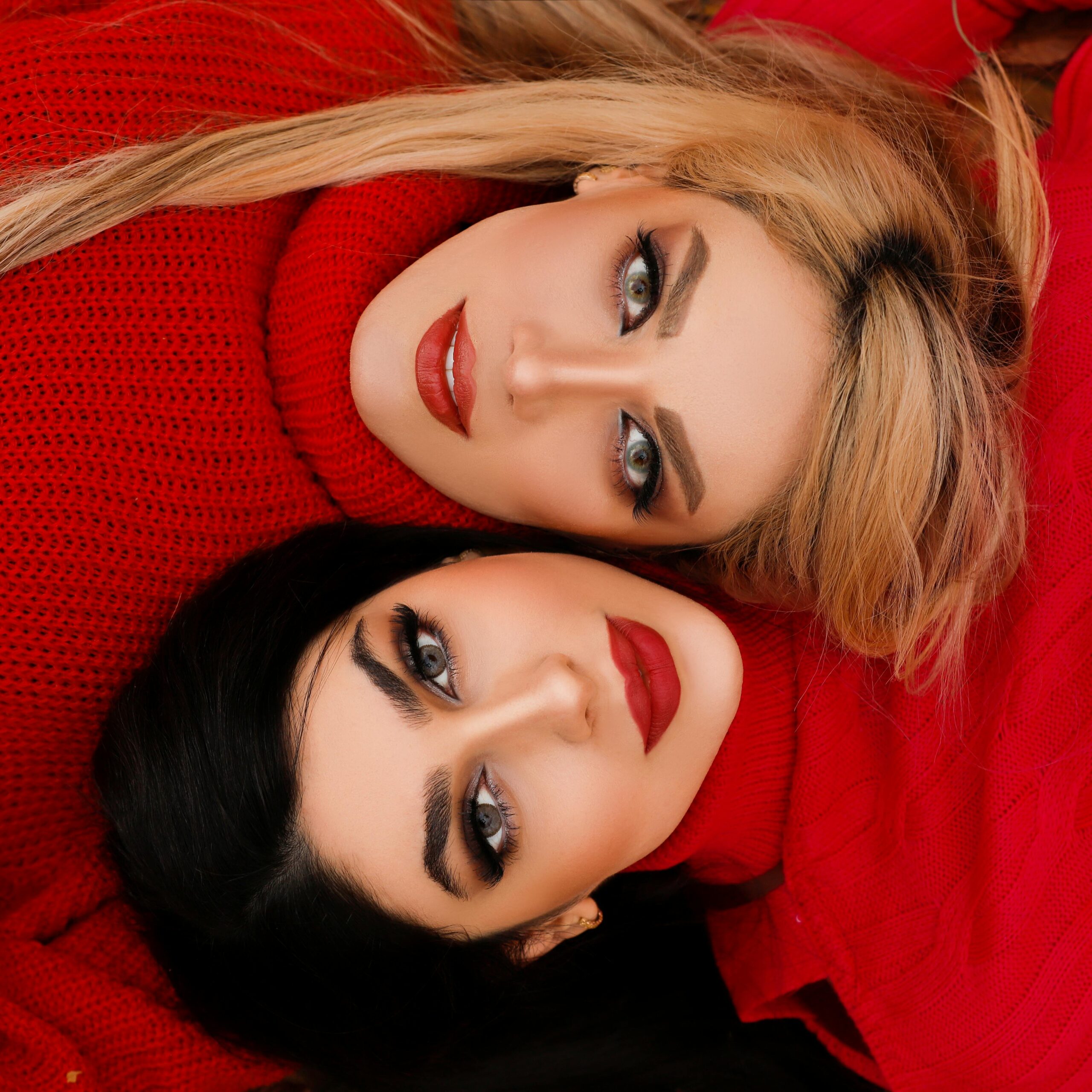 Stylish artistic portrait of two women wearing red sweaters, lying together with striking makeup.