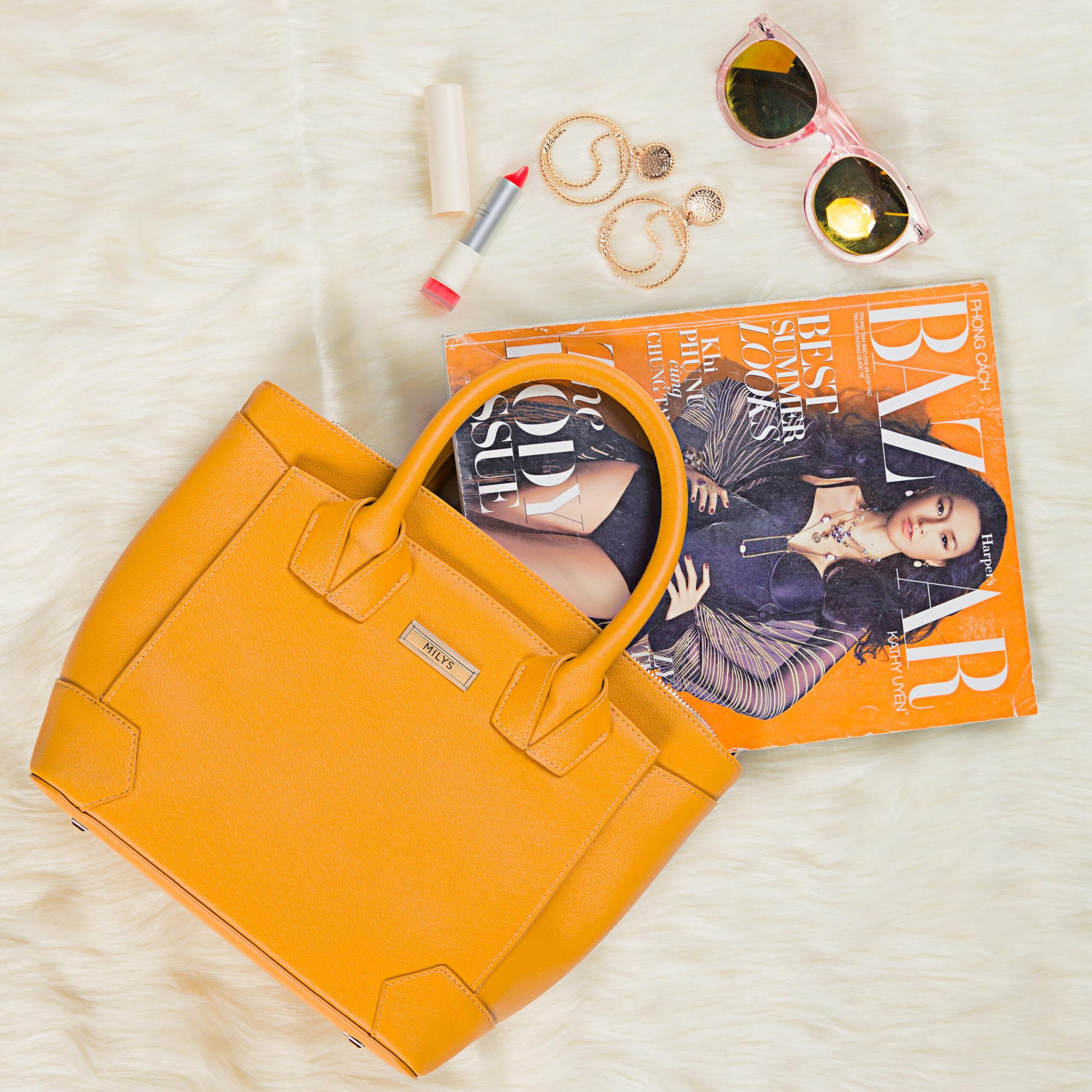 Chic flat lay featuring an orange handbag, sunglasses, lipstick, jewelry, and a fashion magazine.