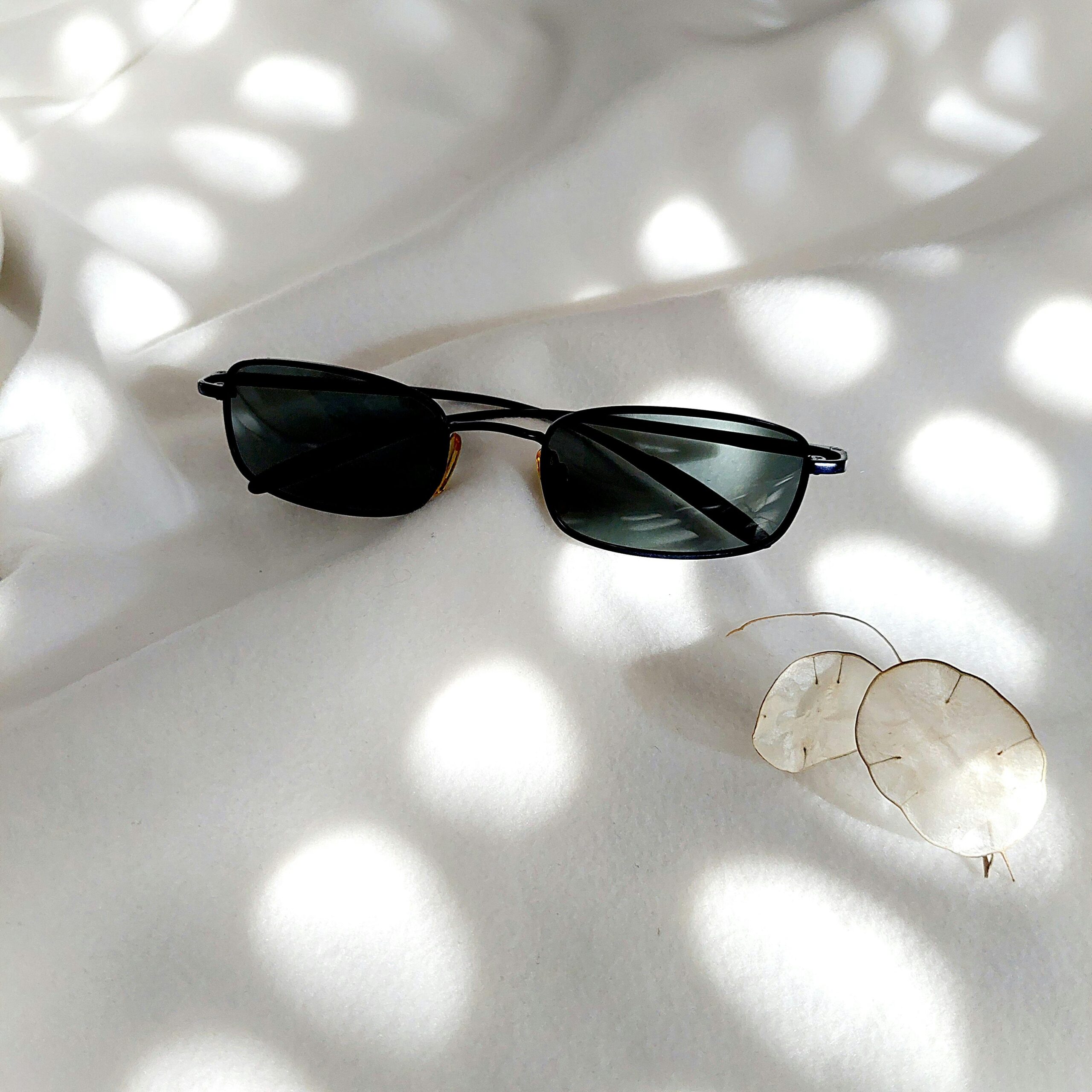 Modern sunglasses rest on a soft white surface with delicate dried plant and light patterns, creating a calm minimalist aesthetic.