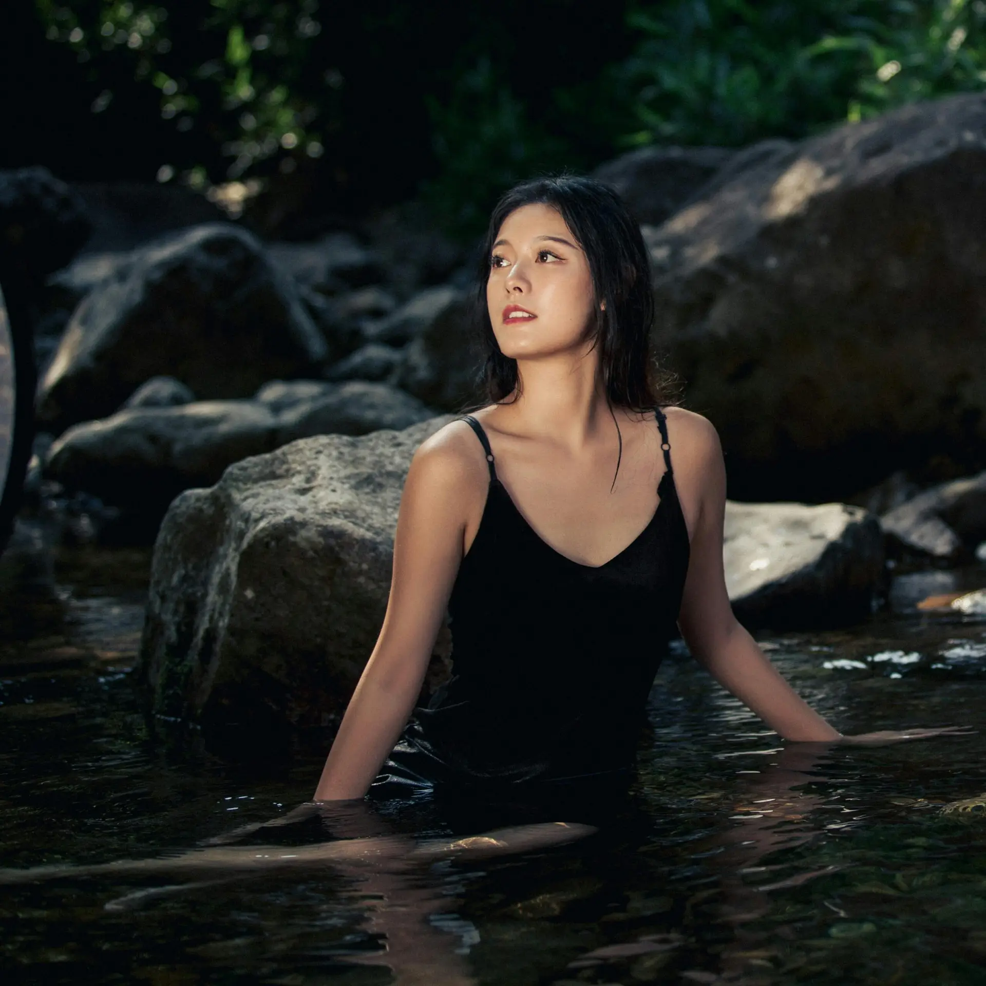 A contemplative woman in a black dress sits in a tranquil rock pool, surrounded by nature.