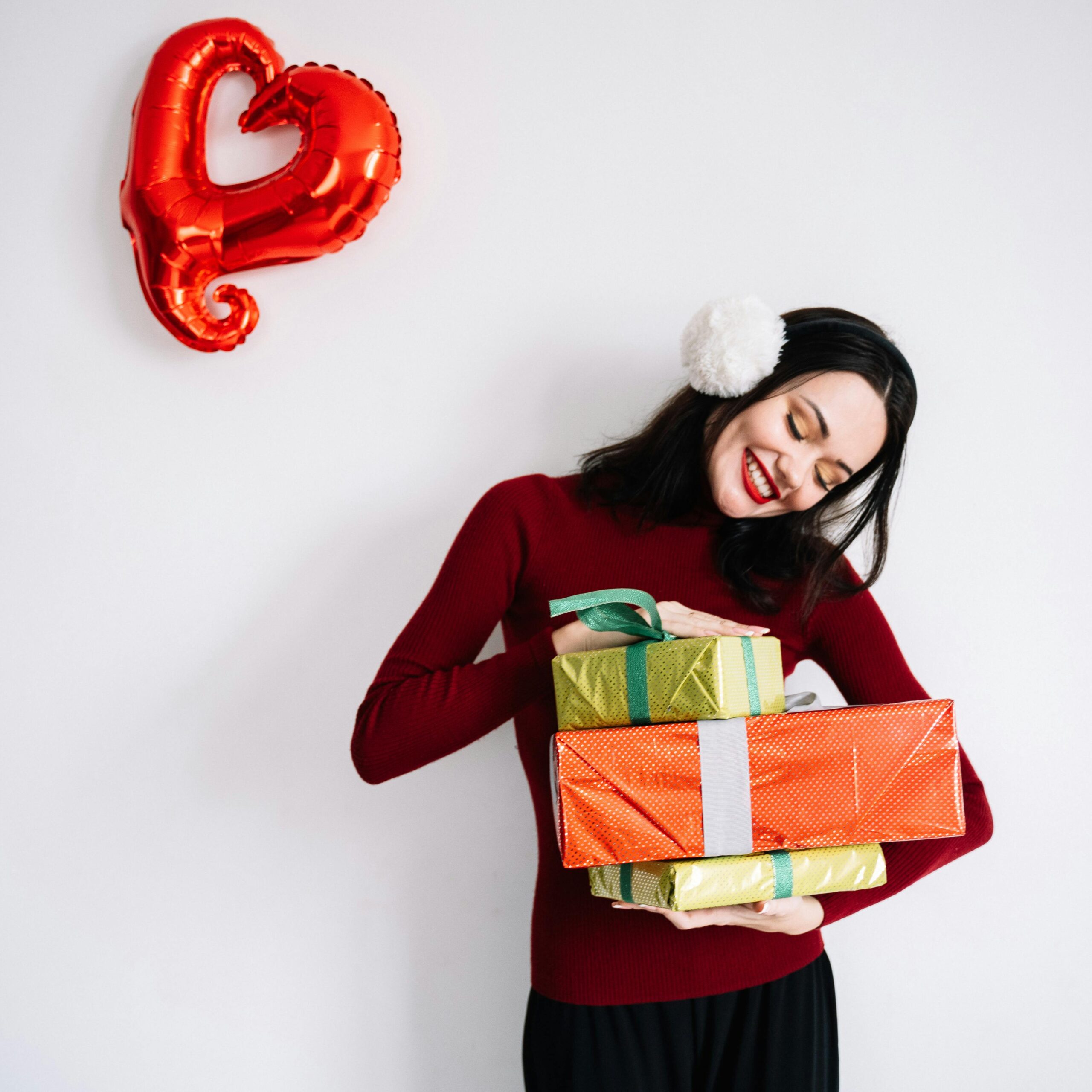 Smiling woman in red holding gift boxes beside heart-shaped balloon.