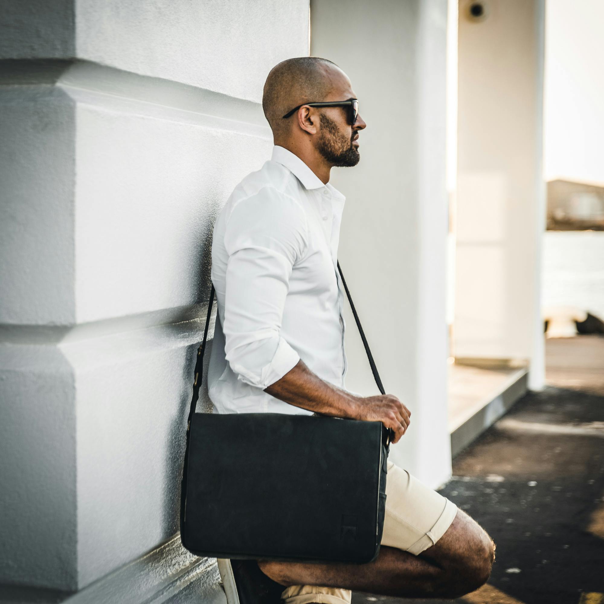 A fashionable man with sunglasses and a bag leans against a wall in an outdoor setting.
