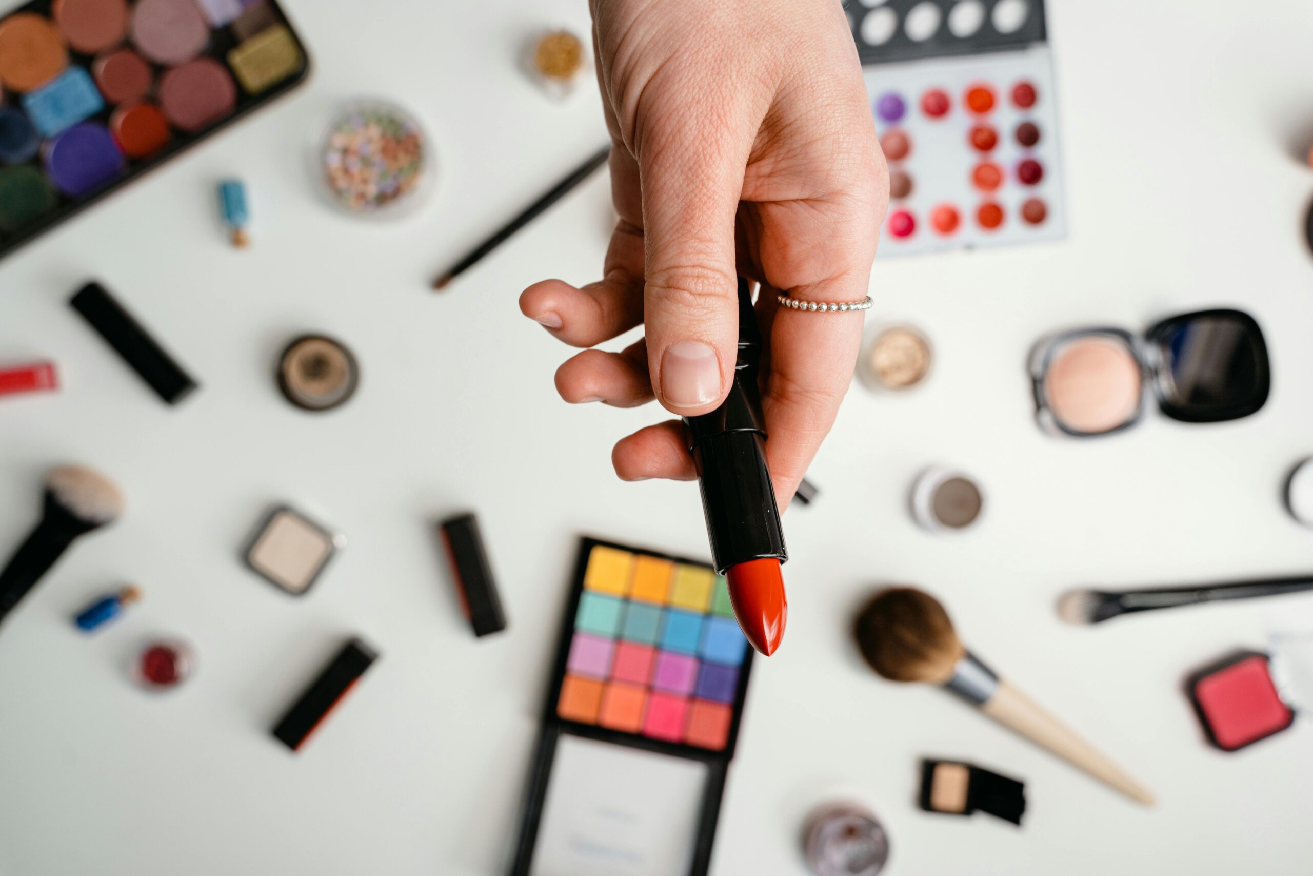 A selection of makeup products with a focus on a vibrant red lipstick held in hand.