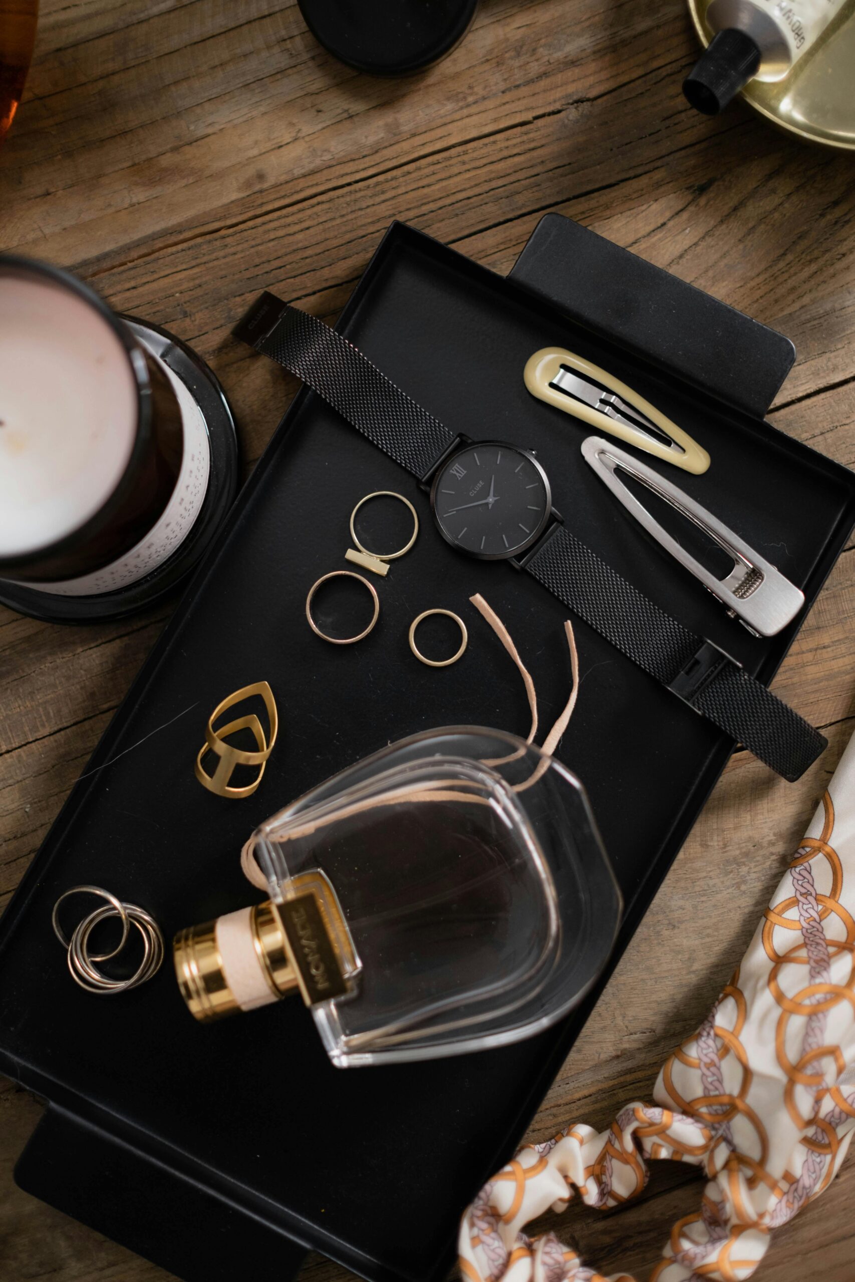 A stylish flatlay featuring a watch, perfume bottle, hair clips, rings, and accessories arranged on a black tray.