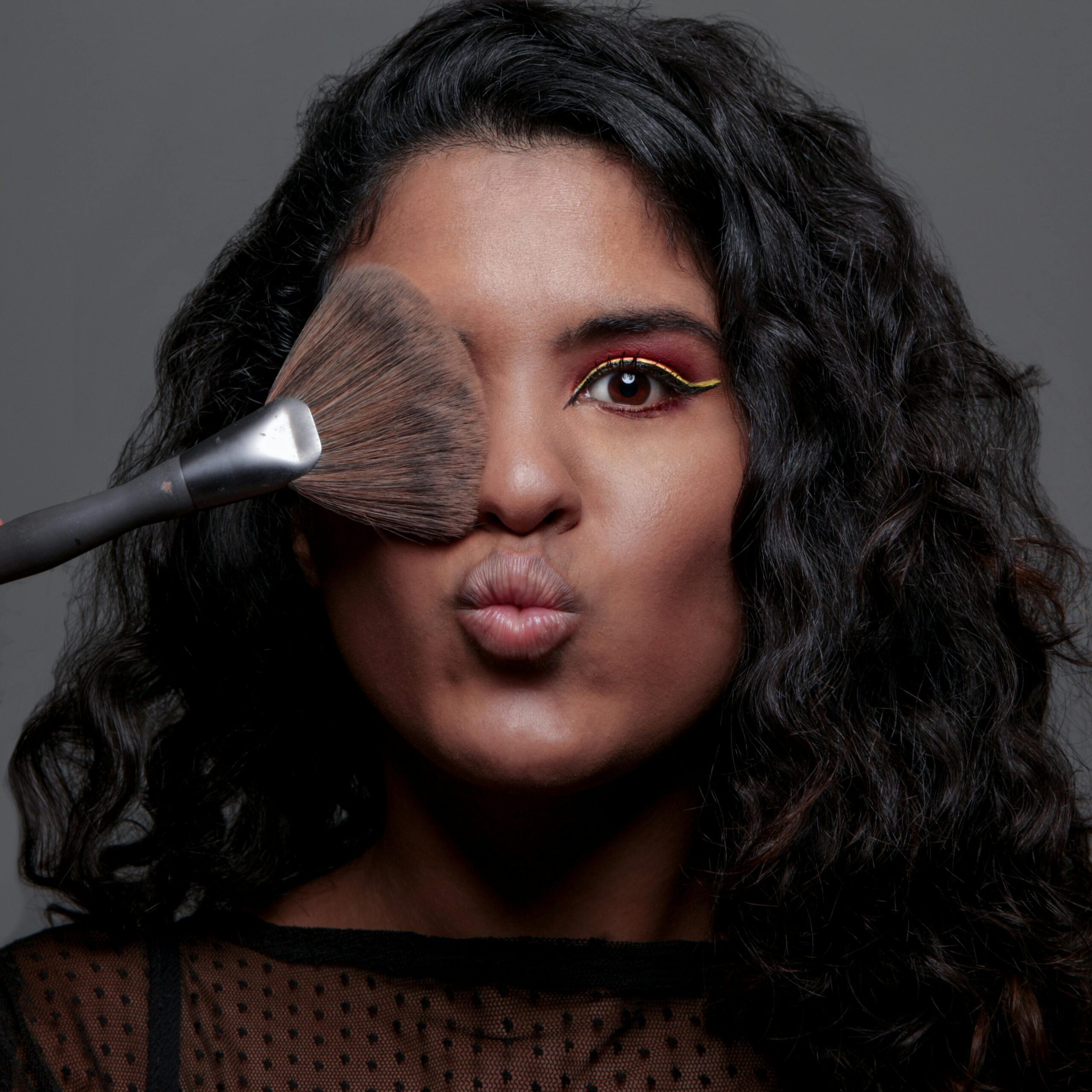 Close-up portrait of a woman with vibrant eye makeup, using a brush, and making a playful expression.