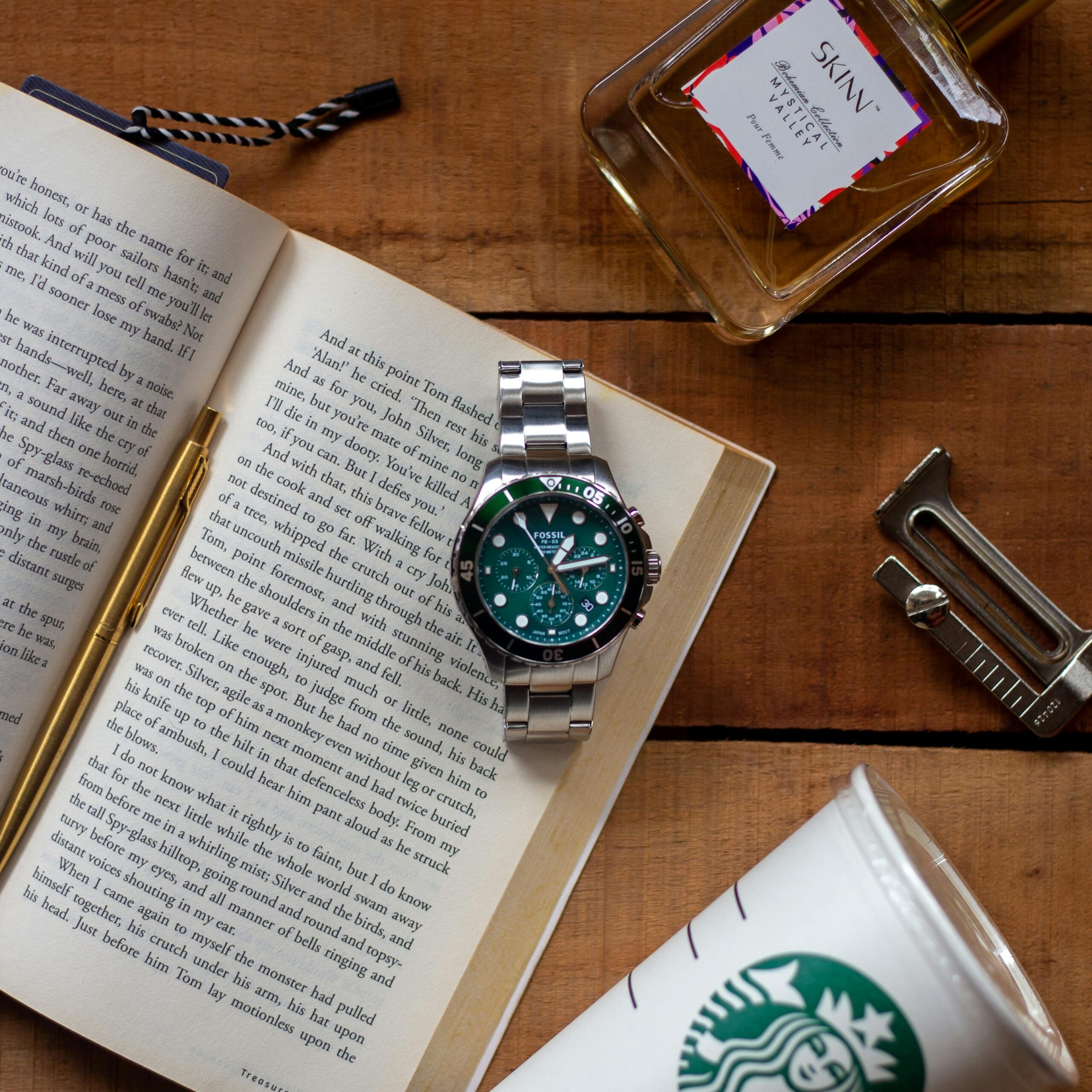 Elegant flat lay featuring a watch on an open book with a perfume bottle and coffee cup on a wooden table.