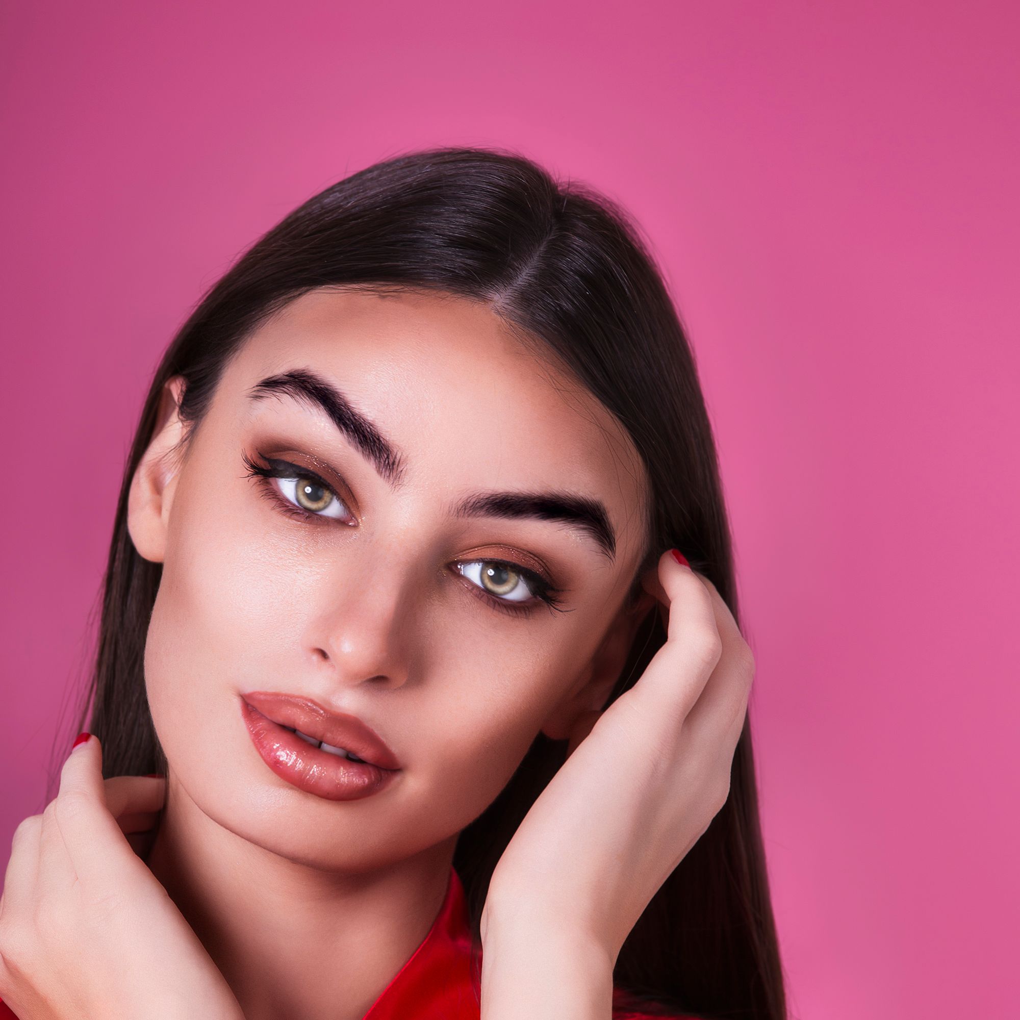Close-up portrait of a woman with striking eyes and long hair against a vibrant pink background.