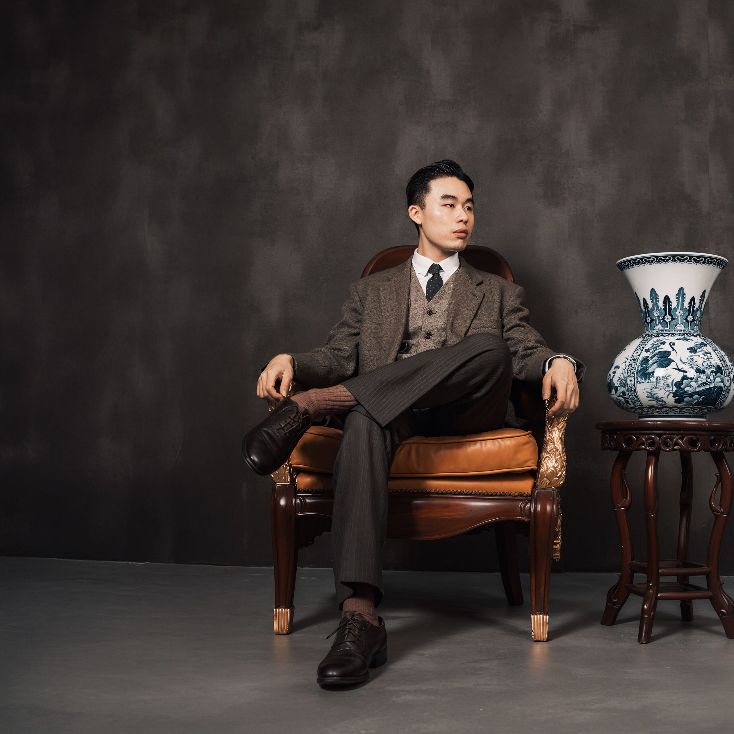 A sophisticated man sits in a classic armchair beside a decorative porcelain vase.