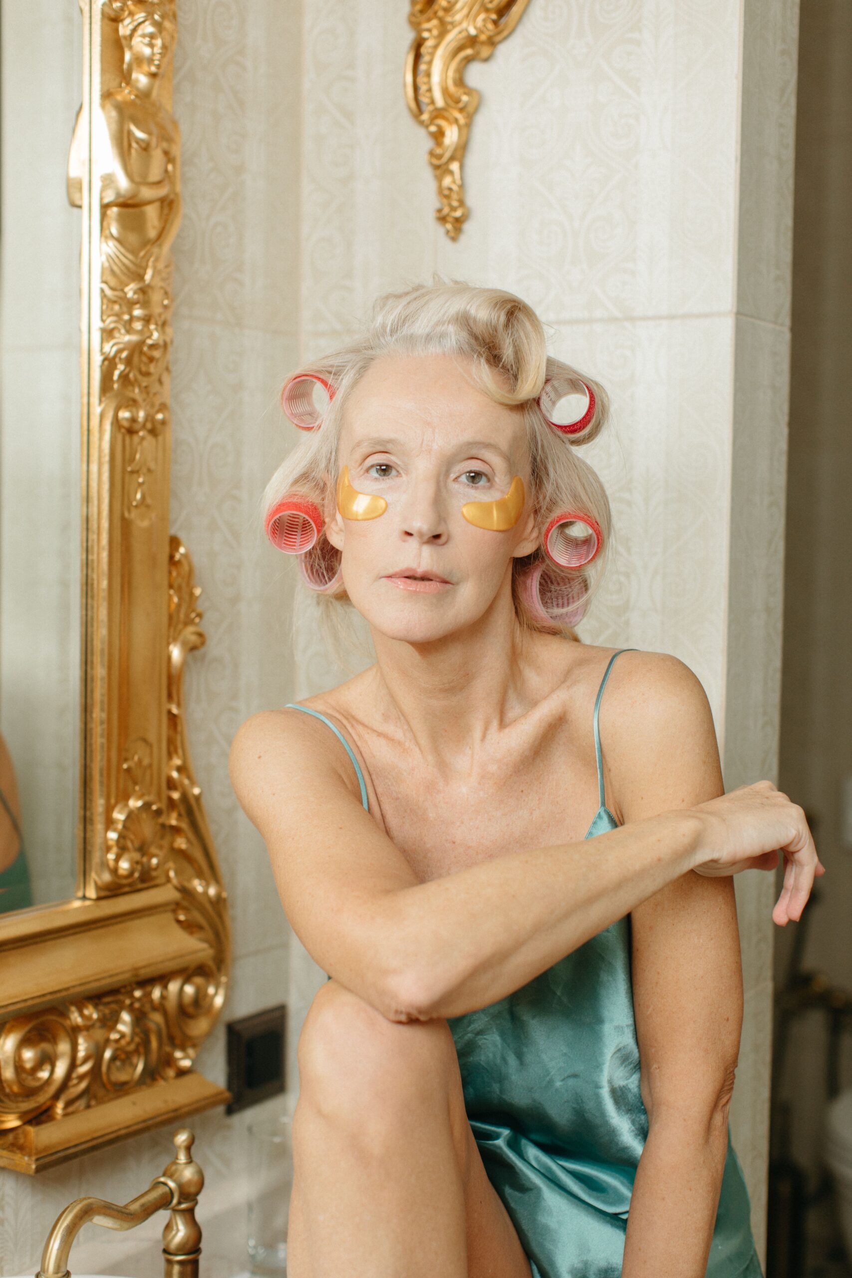A senior woman in hair rollers practicing her skincare routine, sitting elegantly in a vintage bathroom.